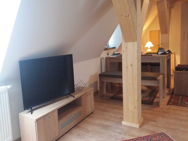 A loft-style room with a flat-screen TV on a wooden cabinet, a dining area with a table and chairs, and a lamp on a desk.