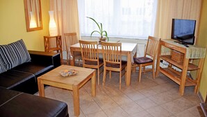A living room with a sofa, a coffee table, a dining table with chairs, a TV, and a window with curtains. - Ferienappartement 05 - Pension in Ostseebad Sellin (Sellin)