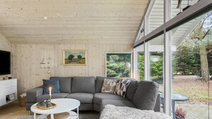 Een moderne woonkamer met een grijze modulaire bank, een witte salontafel en een groot raam met uitzicht op een bebost gebied.