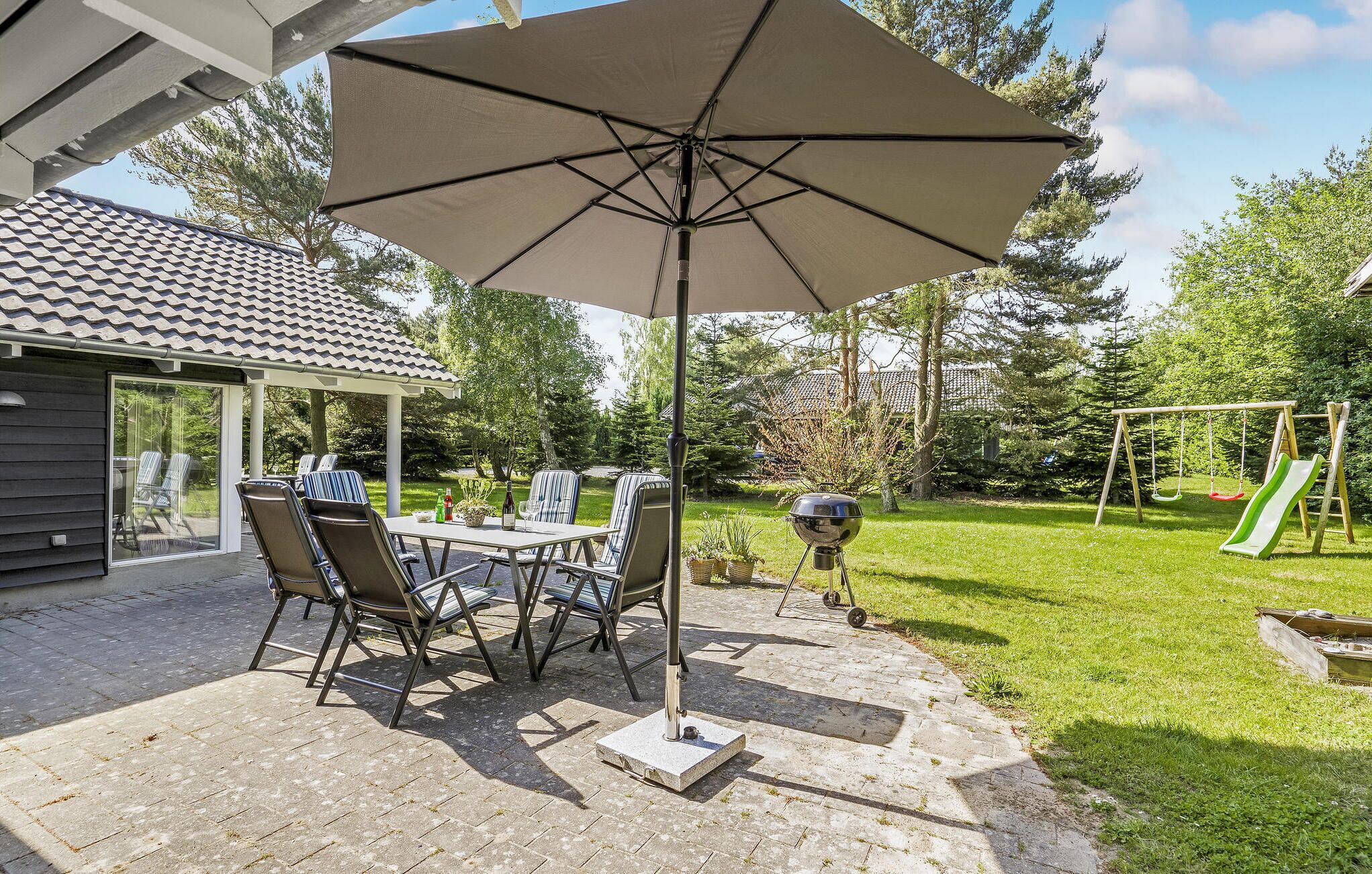 Een terras met een tafel, stoelen en een grote parasol, een barbecue en een schommelset in een achtertuin.