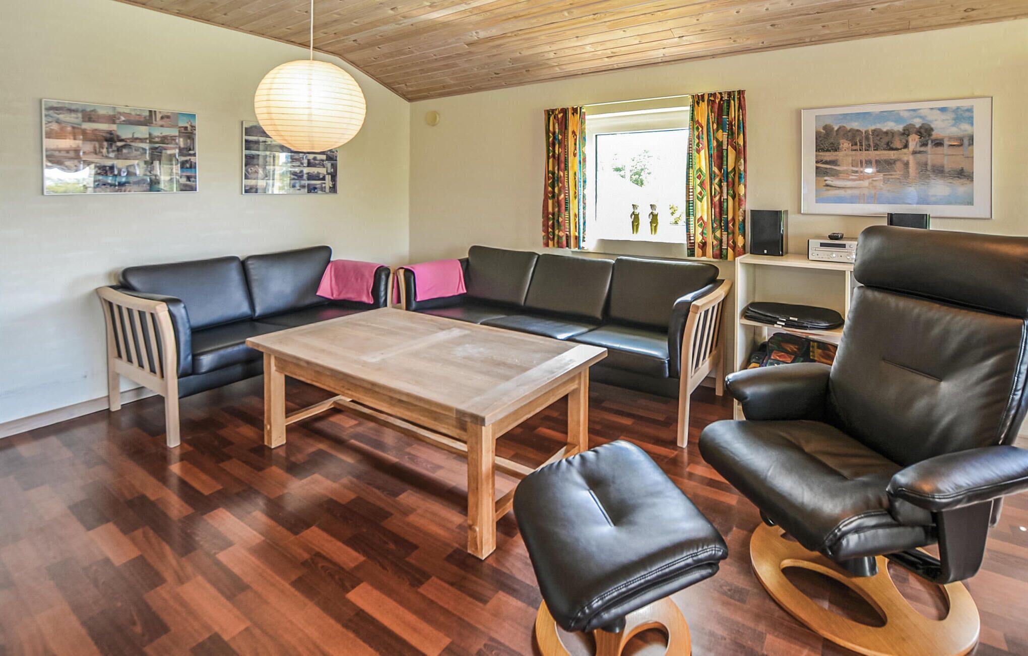 A living room with a wooden table, black leather sofa, and a leather armchair. There are two windows with colorful curtains, a wooden ceiling, and wall-mounted artwork.