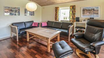 A living room with a wooden table, black leather sofa, and a leather armchair. There are two windows with colorful curtains, a wooden ceiling, and wall-mounted artwork.