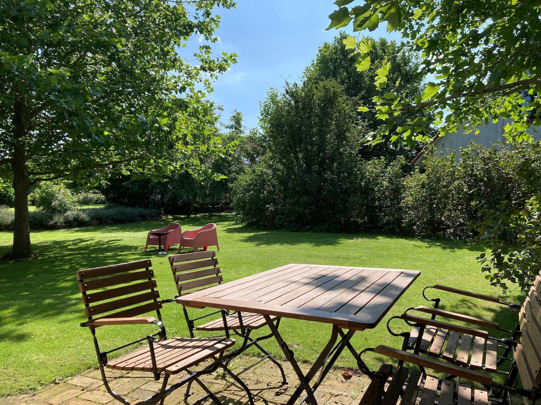 A patio with a wooden table and chairs, surrounded by lush greenery and trees.