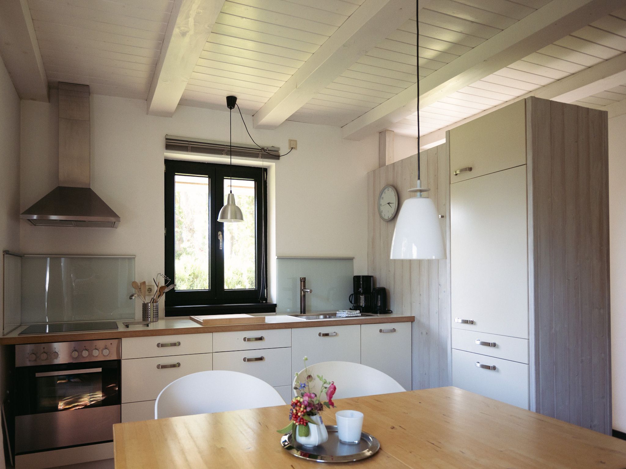 A modern kitchen with a wooden table, white chairs, a stainless steel oven, and a window with a view of greenery.