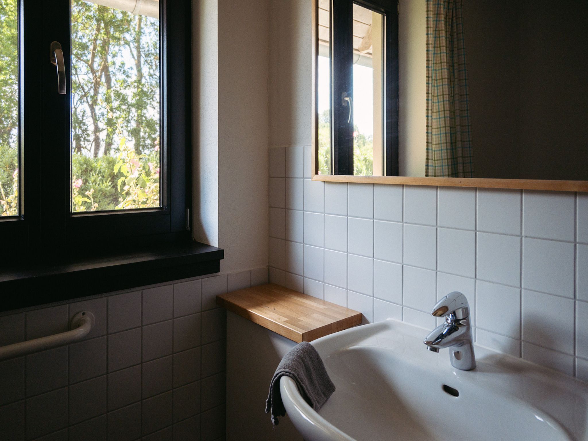 Une salle de bain avec un lavabo blanc, une étagère en bois, une fenêtre donnant sur des arbres et un miroir.