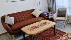 En stue med en brun sofa, et lille træbord, en stol, et billede i ramme på væggen og en potteplante.