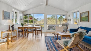 A spacious room with a high ceiling, wooden flooring, a blue sofa, a dining table with chairs, and large windows offering a view of the outdoors. - Lovely home in ålbæk with WiFi (Ålbæk)