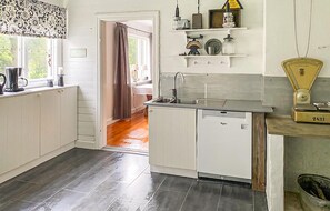 Une cuisine avec des meubles blancs, un lave-vaisselle et un évier. Il y a une fenêtre avec un rideau, et une étagère avec divers objets au-dessus du plan de travail.