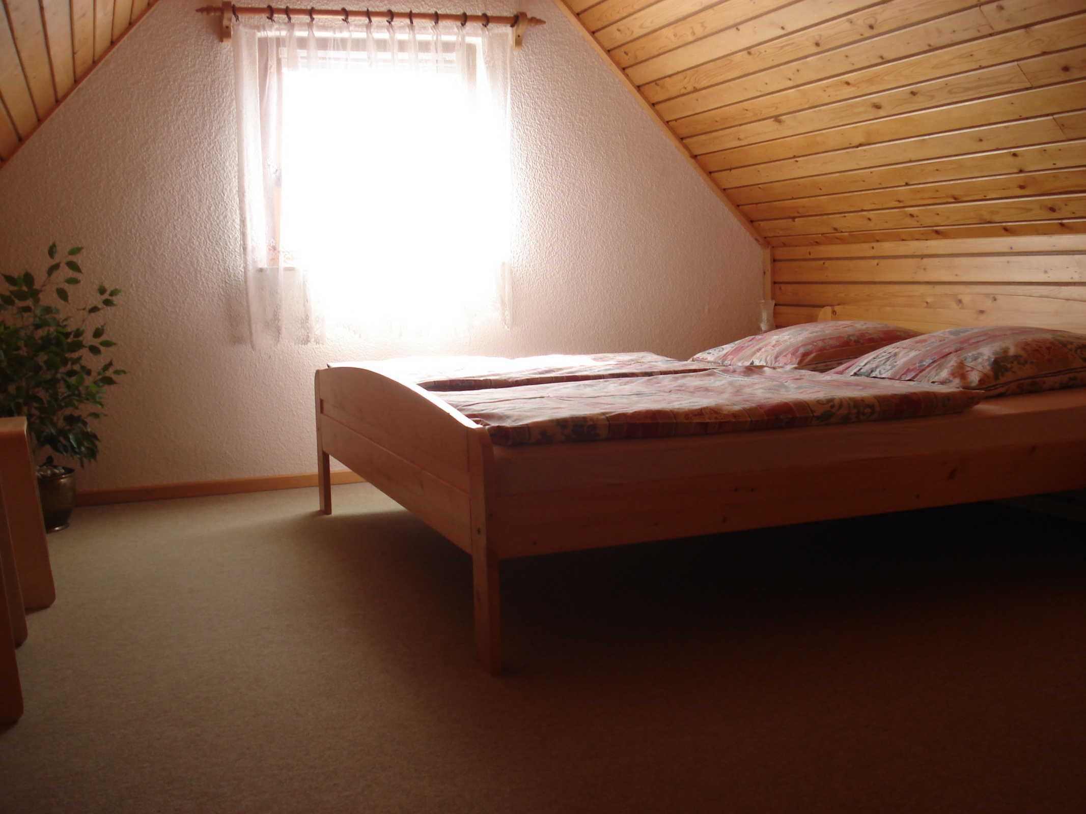 Een houten bed in een kamer met een schuine balkenkap en een raam met gordijn.