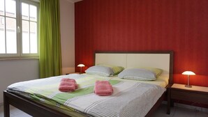 A neatly made bed with striped bedding and two pink towels, a bedside table with a lamp, and a window with green curtains.