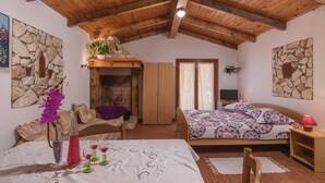 Ein gemütliches Schlafzimmer mit Kamin aus Stein, Holzbalkendecke, einem Bett mit gemusterten Betttextilien, einem kleinen Tisch mit einer Vase und Gläsern sowie einer Steinwand mit Kunstwerken.