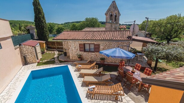 Ein Poolbereich mit Liegestühlen und einem Tisch, ein Steinhaus mit Glockenturm und Blick auf die umliegende Grünfläche.