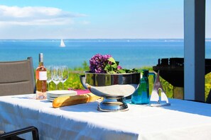 Ein gedeckter Tisch mit einer Weinflasche, Gläsern, einem Brotlaib, einer Metallschale mit Blumen und einem Segelboot im Hintergrund.