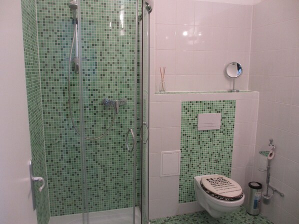 A bathroom with green mosaic tiles, a glass-enclosed shower, a toilet with a decorative cover, and a small mirror on the wall. - Pension Domke Garni - alte Post (Heringsdorf)