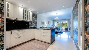 Cocina moderna con armarios blancos, encimera negra y suelo de madera. Hay una zona de comedor con mobiliario azul y vistas a zonas verdes.