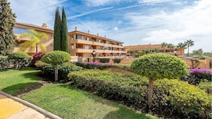 Un hotel con un patio que cuenta con setos cuidados, una palmera y un cielo azul despejado.