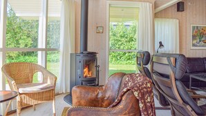 A cozy room with a wood-burning stove, wicker chairs, a leather sofa, and large windows offering a view of greenery. - Stunning home in Gilleleje with kitchen (Gilleleje)