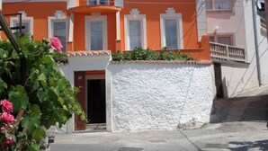 A two-story building with a white and orange facade, a balcony, and a small garden area with pink flowers.