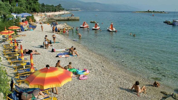 A pebbled beach with umbrellas and sun loungers, boats in the water, and people swimming and relaxing.
