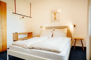 Una cama bien hecha con ropa de cama blanca, una mesita de noche de madera, un estante en la pared y un cuadro enmarcado sobre la cama.