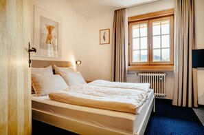 A hotel room with a bed, a window with curtains, a radiator, and a framed picture on the wall. - Basic double rooms (16m²) - Hotel & Restaurant Alpenrose Bayrischzell (Bayrischzell)