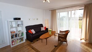 Ein Wohnzimmer mit einer schwarzen Couch, einem weißen Bücherregal, einem Holztisch und einem Korbstuhl.
