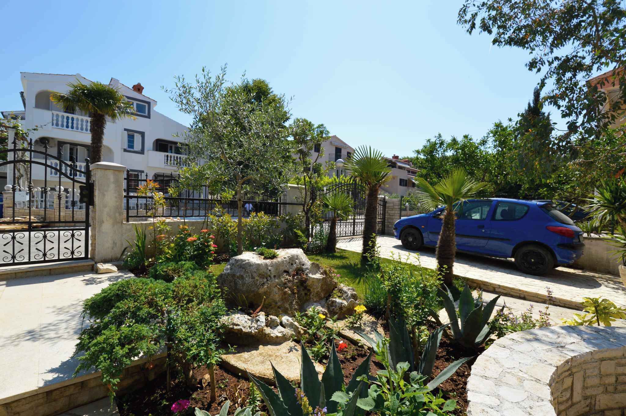A residential area with a blue car parked on the street, a white building with balconies, and a garden with various plants and a rock centerpiece.