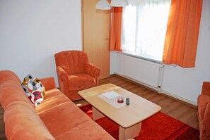 A room with a wooden floor, a sofa, a chair, a small table with a remote control, and a window with orange curtains.