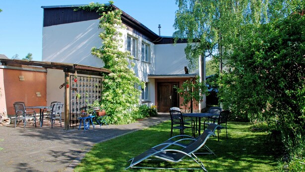 A building with a white exterior, a wooden door, and a small garden area with chairs and a table. - Apartment OG - Apartments on the Heideweg (Baabe)
