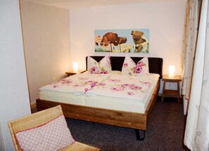 A bedroom with a wooden bed, floral bedding, two bedside lamps, a painting on the wall, and a chair with a patterned cushion.
