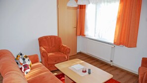 A room with a wooden floor, a sofa, a chair, a small table with a remote control, and a window with orange curtains. - Apartment OG - Apartments on the Heideweg (Baabe)