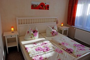 Un dormitorio con una cama blanca, ropa de cama floral, dos mesitas de noche con velas y una ventana con cortinas.