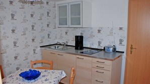 A compact kitchen with a sink, a coffee maker, and a microwave. There is a dining table with a blue floral tablecloth and a blue bowl. The walls have a pattern of coffee-related icons. - Apartment OG - Apartments on the Heideweg (Baabe)
