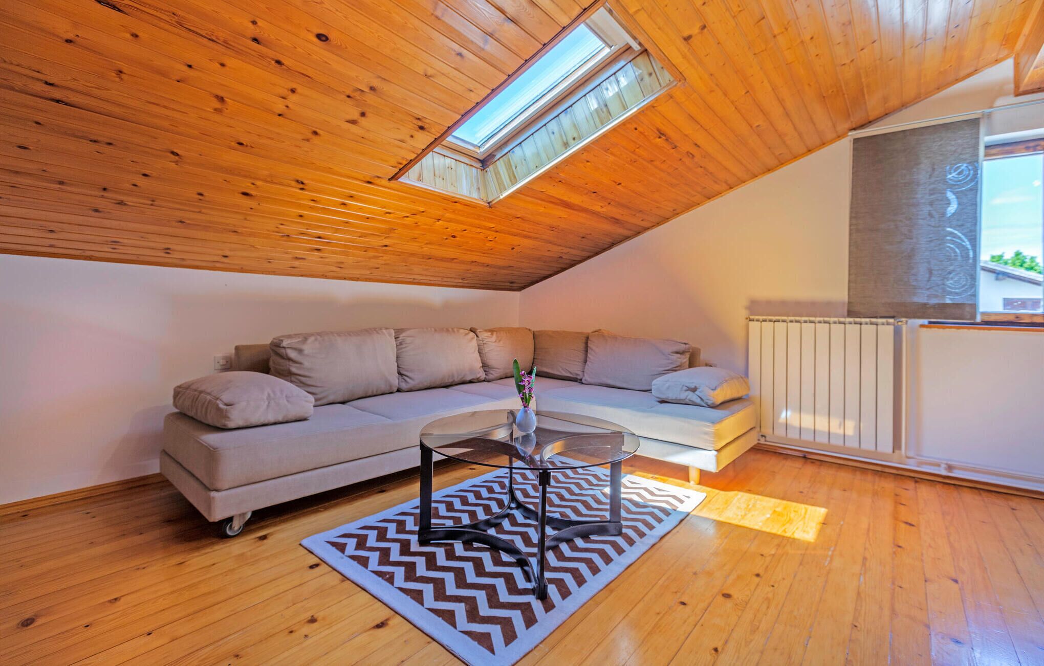 A modern loft bedroom with a wooden ceiling, a large window, a sofa, a glass coffee table, and a radiator.