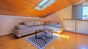 A modern loft bedroom with a wooden ceiling, a large window, a sofa, a glass coffee table, and a radiator.