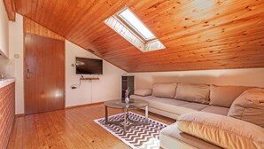 A cozy loft bedroom with a wooden ceiling, a large window, a TV, a sofa, and a small table.