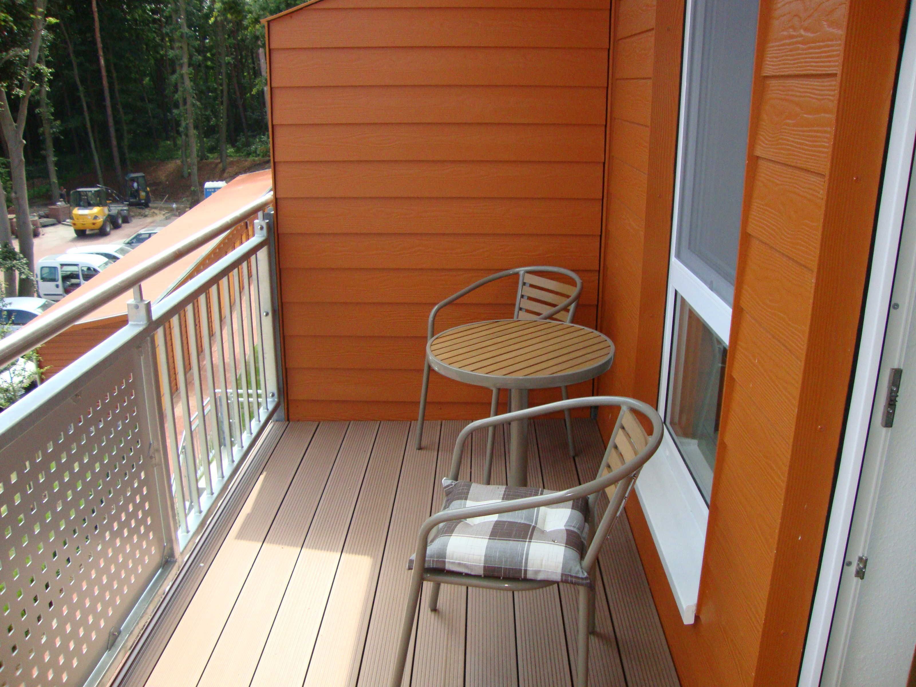 A balcony with a wooden table and two chairs, a metal railing, and a view of the street with parked cars.