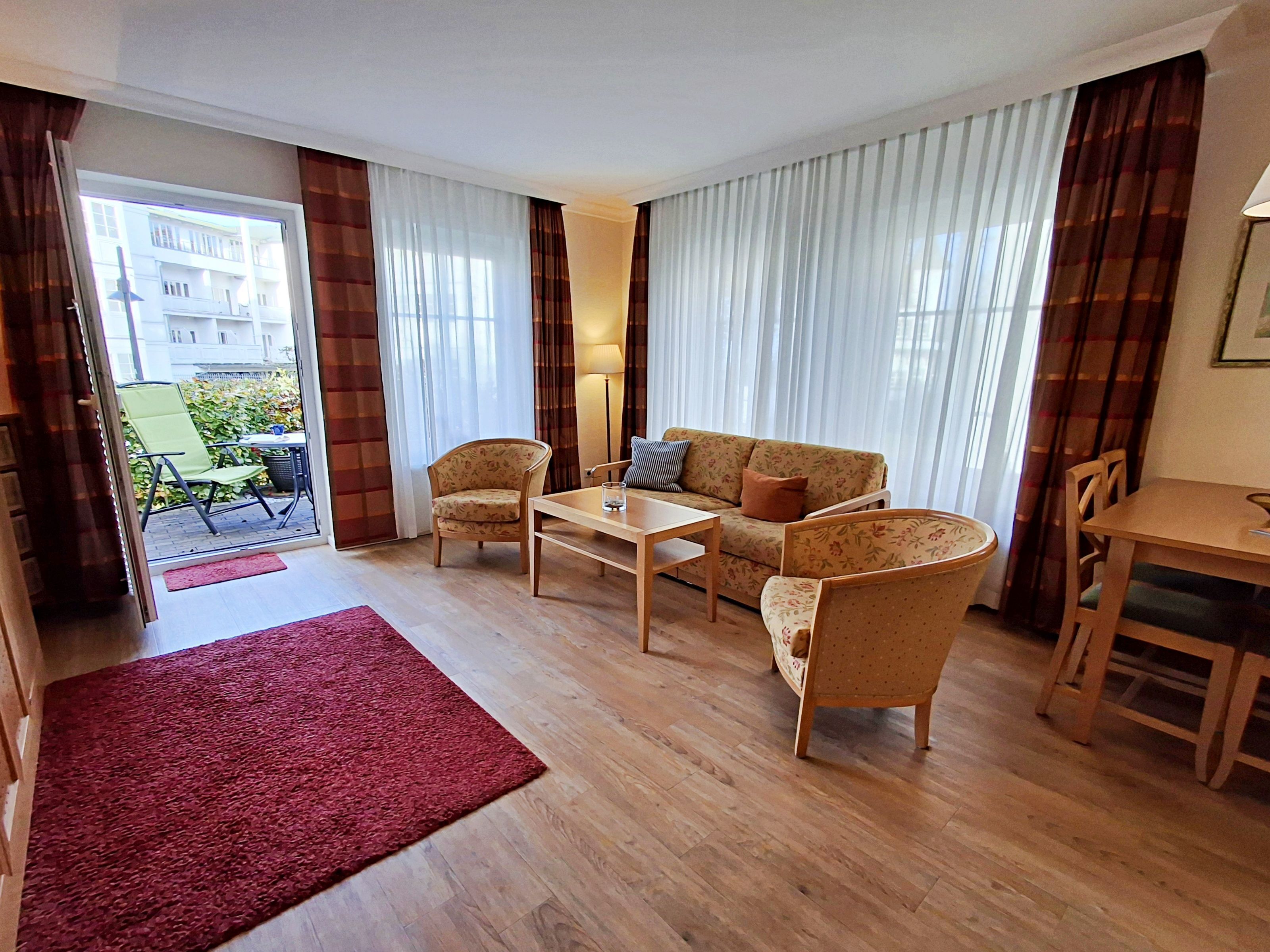 A living room with a sofa, two armchairs, a coffee table, and a dining table. There is a red rug on the floor, and the room has large windows with curtains.
