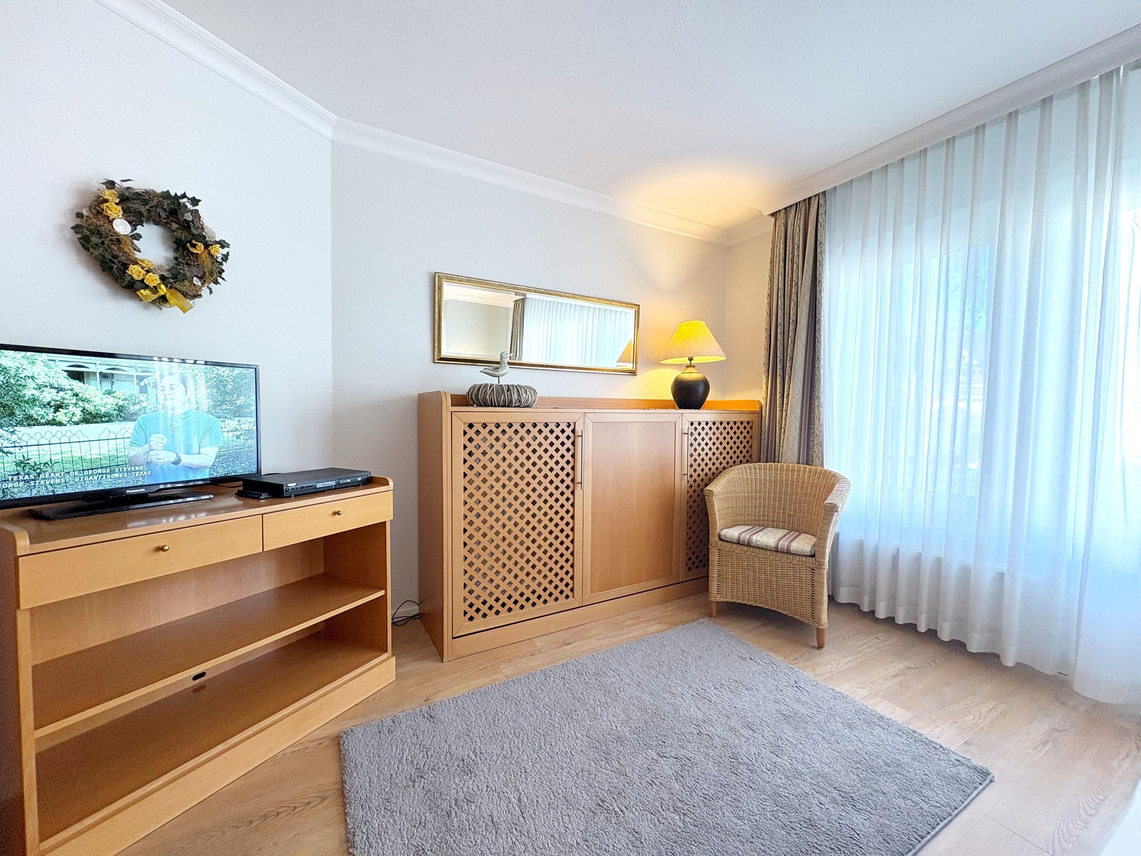 A hotel room with a flat-screen TV, wooden furniture, a wicker chair, a mirror, and sheer curtains.