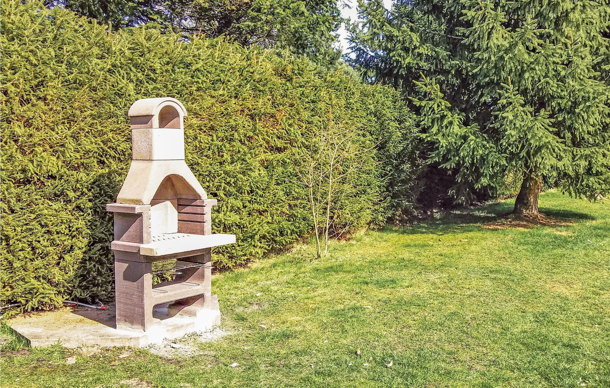 A stone barbecue grill with a chimney, set on a concrete base, surrounded by a well-maintained lawn and dense shrubbery.