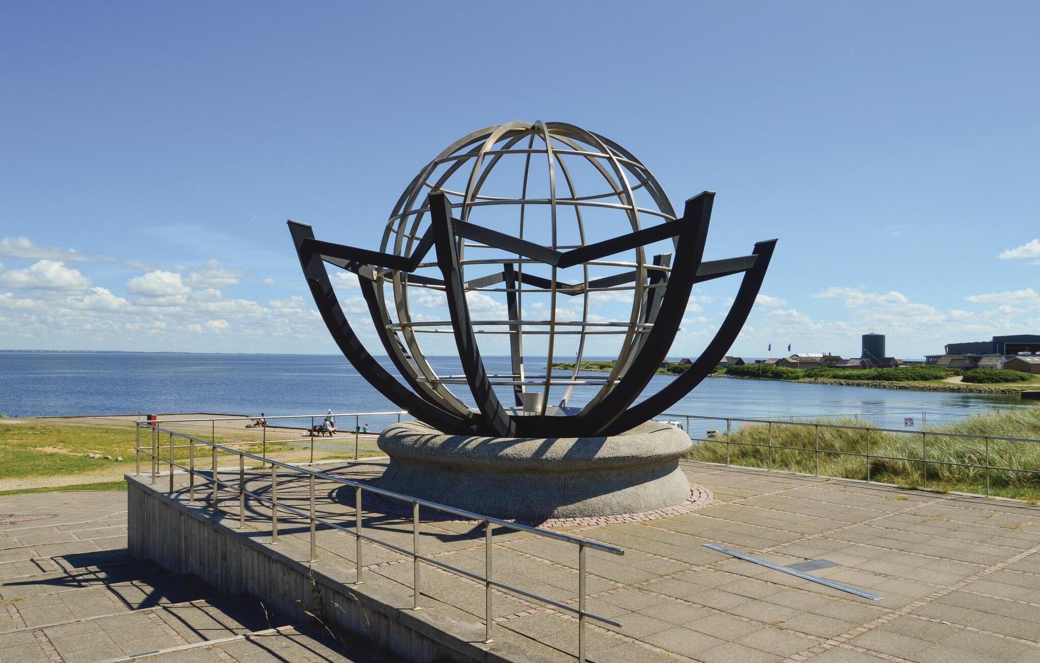 Uma grande escultura de metal que se assemelha a um globo, com uma base, localizada em área pavimentada com vista para o mar e edifícios ao fundo.