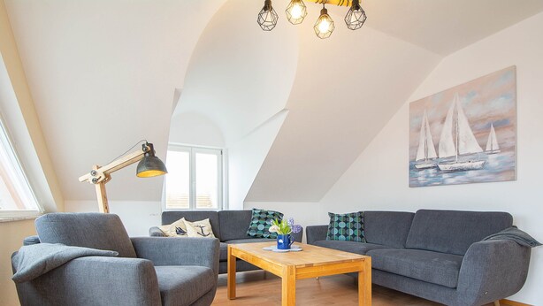 Una sala moderna con un sofá gris, un sillón gris y una mesa de centro de madera. Hay un cuadro de veleros en la pared y una lámpara con pantalla geométrica.