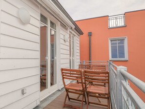 Un balcon avec des chaises et une table en bois, des murs recouverts d’un bardage blanc, et un bâtiment rouge en arrière-plan.