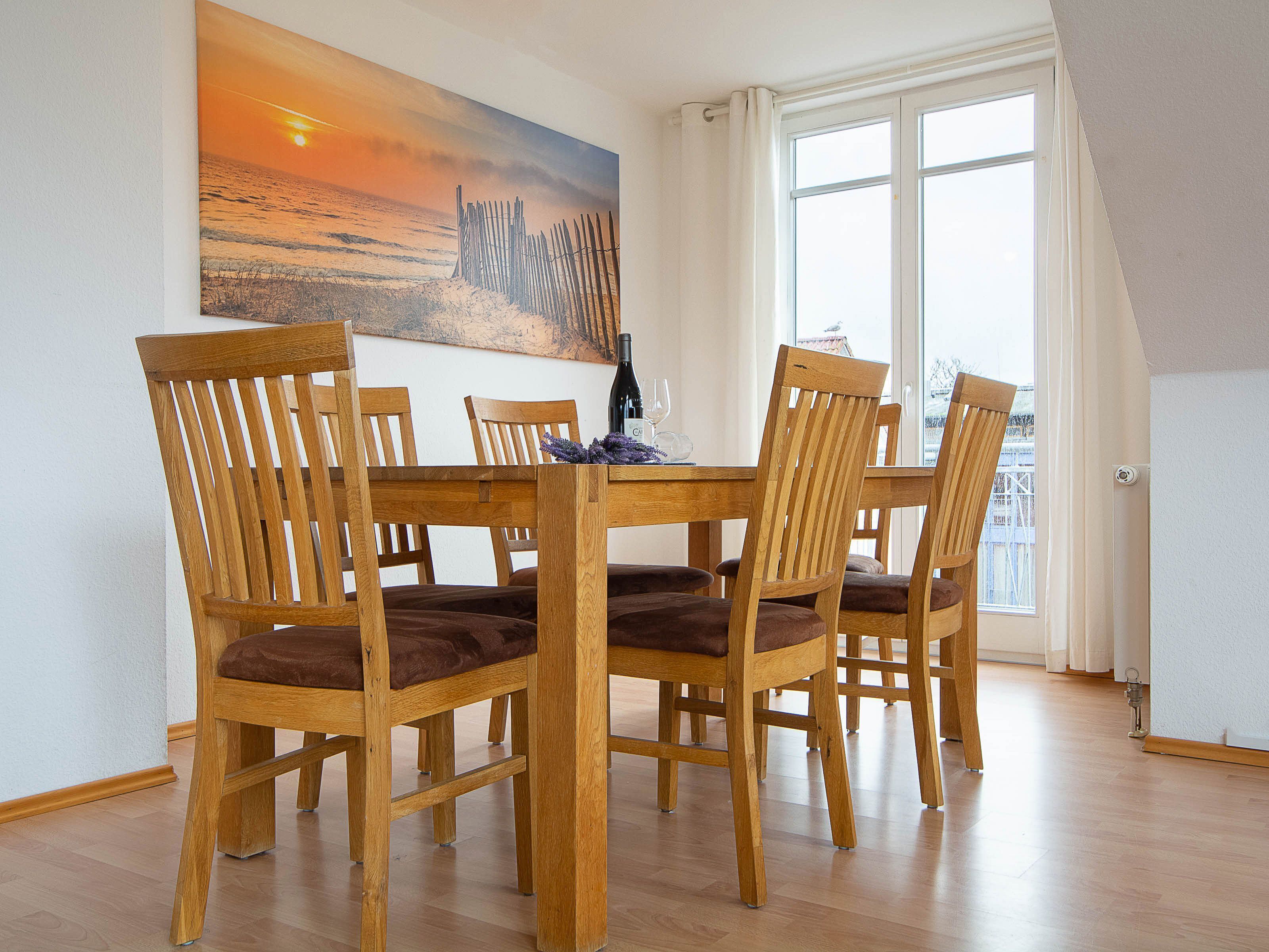 A dining area with a wooden table and chairs, a painting on the wall, and a view through a window.