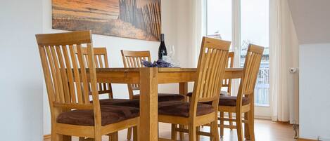 A dining area with a wooden table and chairs, a painting on the wall, and a view through a window.