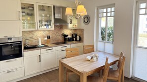 A modern kitchen with white cabinets, a built-in oven, a sink, and a dining area with a wooden table and chairs. - Apartment 02 with fireplace & terrace - A: Haus Strandkiefer - close to the beach (Baabe)