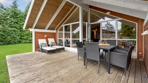 Een overdekt terras met een houten vloer, twee loungesteunen, een eettafel met stoelen en grote ramen.