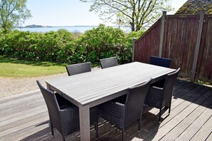 Una terraza de madera al aire libre con una mesa y sillas, rodeada de follaje y vistas a un cuerpo de agua.