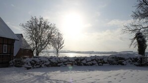 A snow-covered landscape with a house, a stone wall, and a body of water in the background.