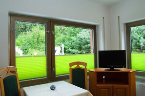 Quarto com televisão sobre suporte de madeira, mesa com toalha branca, duas cadeiras e janelas amplas com persianas.